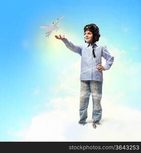 Image of little boy in pilots helmet playing with toy airplane against clouds background