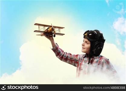 Image of little boy in pilots helmet playing with toy airplane against clouds background