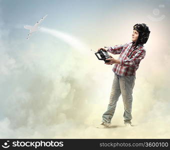 Image of little boy in pilots helmet playing with toy radiocontrolled airplane against clouds background