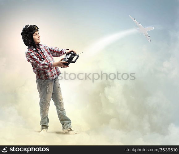 Image of little boy in pilots helmet playing with toy radiocontrolled airplane against clouds background