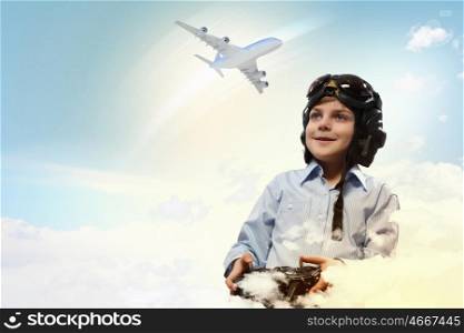 Image of little boy in pilots helmet playing with toy radiocontrolled airplane against clouds background