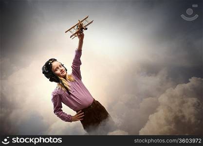Image of little girl in pilots helmet playing with toy airplane against clouds background