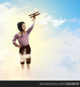 Image of little girl in pilots helmet playing with toy airplane against clouds background