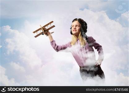 Image of little girl in pilots helmet playing with toy airplane against clouds background