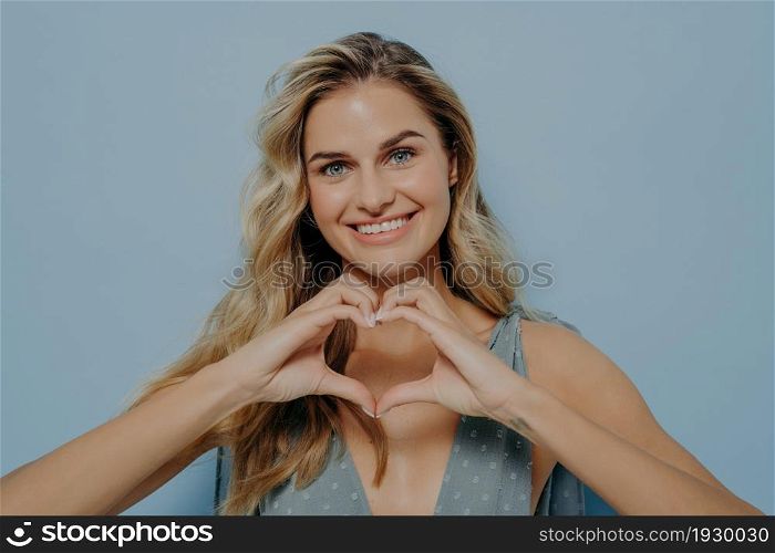 Image of lovely blonde woman in blue dress admitting in her love with use of heart gesture she made out of her hands, holding it in front while standing isolated next to blue background. Lovely blonde woman admitting in her love with use of heart gesture