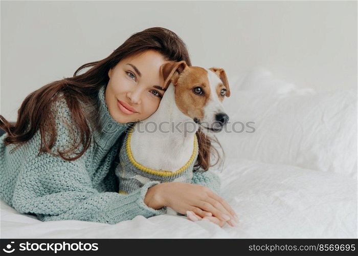 Image of lovely brunette lady leans at pedigree jack russel terrier, embraces pet with love, spend time together, lie on white bedclothes. People, animals, love and friendship concept. Dog lover