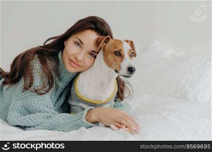 Image of lovely brunette lady leans at pedigree jack russel terrier, embraces pet with love, spend time together, lie on white bedclothes. People, animals, love and friendship concept. Dog lover