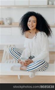 Image of lovely curly haired lady drinks coffee or tea from white mug, wears fashionable white sweater, striped pants, poses at kitchen against blurred background. People and lifestyle concept