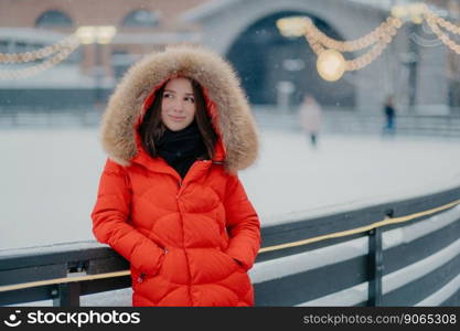 Image of lovely thoughtful young woman wears winter red coat with fur on hoody, keeps hands in pockets, stands against blurred background near skating rink, has outdoor walk during winter time