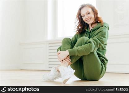 Image of satisfied refreshed young woman with foxy hair, keeps legs crossed, wears tracksuit and sportshoes, glad to have workout, sits on floor in spacious empty hall for doing sport exercises