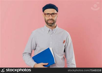 Image of self confident young male with stubble, wears transparent glasses, holds textbook under arm, prepares project work, models against pink background. Businessman with notepad poses indoor.