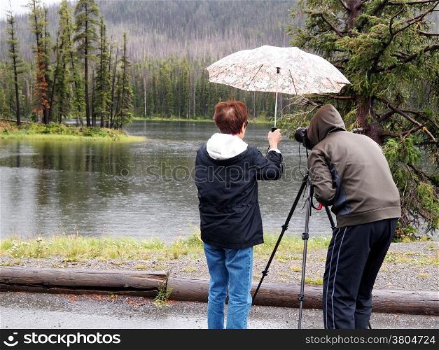Image of senior women holding umbrella over photographer head while taking photos of small lake in pouring rain