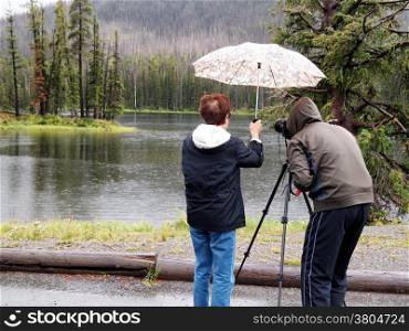 Image of senior women holding umbrella over photographer head while taking photos of small lake in pouring rain