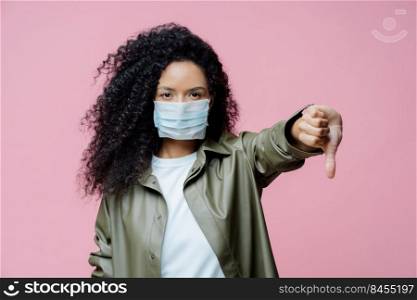 Image of serious curly haired young woman wears medical mask, keeps thumb down, shows dislike gesture, avoids spreading of corona virus, cares about health. Dangerous virus. Medical concept.