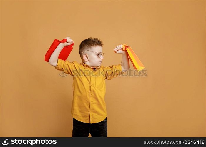 Image of smiling boy holding bags with presents or shoppings on yellow background. child in glasses lifts packages like weights.. Image of smiling boy holding bags with presents or shoppings on yellow background. child in glasses lifts packages like weights