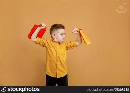 Image of smiling boy holding bags with presents or shoppings on yellow background. child in glasses lifts packages like weights.. Image of smiling boy holding bags with presents or shoppings on yellow background. child in glasses lifts packages like weights