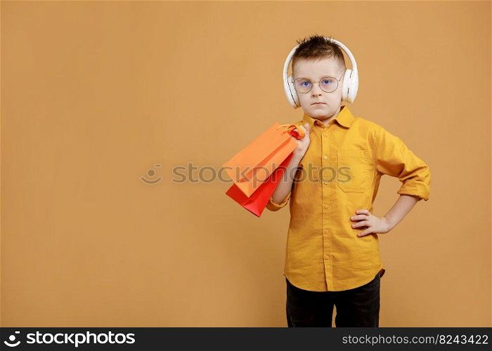 Image of smiling boy holding bags with presents or shoppings on yellow background. child in white headphone. Shopper with many colored paper bags. Holidays sales and discounts. Cyber monday.. Image of smiling boy holding bags with presents or shoppings on yellow background. child in white headphone. Shopper with many colored paper bags. Holidays sales and discounts. Cyber monday