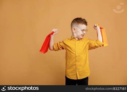 Image of smiling boy holding bags with presents or shoppings on yellow background. child in glasses lifts packages like weights.. Image of smiling boy holding bags with presents or shoppings on yellow background. child in glasses lifts packages like weights