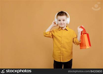 Image of smiling boy holding bags with presents or shoppings on yellow background. child in white headphone. Shopper with many colored paper bags. Holidays sales and discounts. Cyber monday.. Image of smiling boy holding bags with presents or shoppings on yellow background. child in white headphone. Shopper with many colored paper bags. Holidays sales and discounts. Cyber monday