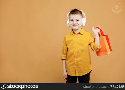 Image of smiling boy holding bags with presents or shoppings on yellow background. child in white headphone. Shopper with many colored paper bags. Holidays sales and discounts. Cyber monday.. Image of smiling boy holding bags with presents or shoppings on yellow background. child in white headphone. Shopper with many colored paper bags. Holidays sales and discounts. Cyber monday