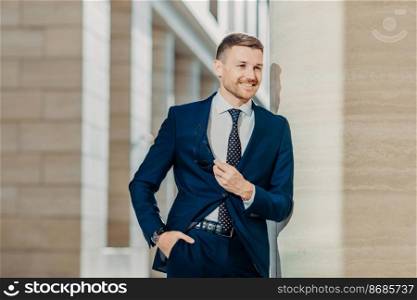 Image of successful businessman in elegant clothing, keeps hand in pocket, holds sunglasses, smiles cheerfully, looks somewhere, being confident in his business deal. People and job concept.
