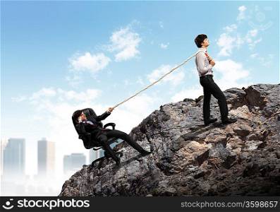 Image of two young businessmen pulling rope atop of mountain against city background