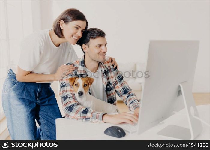 Image of woman touches gently husbands shoulders, look positively in monitor of computer, surf internet, buy furniture online for their new apartment. Happy man works at modern device with dog