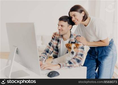 Image of woman touches gently husbands shoulders, look positively in monitor of computer, surf internet, buy furniture online for their new apartment. Happy man works at modern device with dog