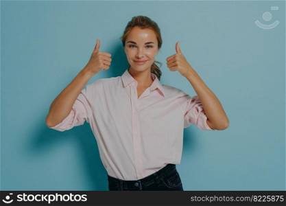 Image of young confident supportive happy young lady shows thumbs up sign with both hands, woman making approval gesture of good quality, standing isolated over blue studio wall background. Young confident supportive happy young lady shows thumbs up sign with both hands