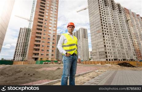 Image of young engineer in hardhat and safety vest posing against buildings under construction