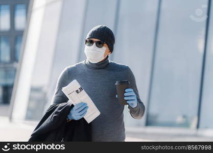 Image of young man wears sterile mask and rubber medical gloves, looks aside, walks through city during infectious disease spread, drinks coffee, folded newspaper. Coronavirus prevention concept