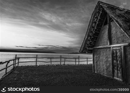 Image with a clay thatched house suspended on stilts over a lake, with the sunset light behind it, in monochrome settings