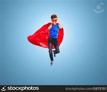 imagination, gesture, childhood, movement and people concept - boy in red super hero cape and mask flying in air and showing thumbs up over blue background