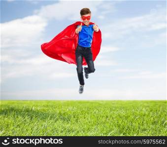 imagination, gesture, childhood, movement and people concept - boy in red super hero cape and mask flying in air and showing thumbs up over blue sky and grass background