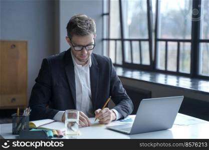Immersed in work. Focused young German male banker writes down memos on sticky notes to remember important details to share for upcoming online meeting while sitting at desk at modern home office. Focused male banker writes down memos on sticky notes while working remotely from home office