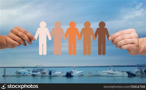 immigration, unity, population, race and humanity concept - multiracial couple hands holding chain of paper people pictogram over boats in sea background