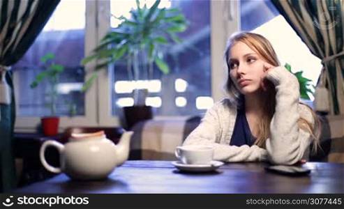 Impatient beautiful young woman waiting for her boyfriend who&acute;s late to the restaurant. Attractive worried teenage girl with blonde hair looking at time on watch, checking out smart phone while sitting alone in coffee shop waiting for tardy boyfriend