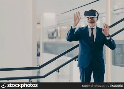 Impressed amazed male office worker wearing portable VR goggles exploring virtual world while standing in office corridor, gesturing with both hands in air, touching objects in digital interface. Impressed male office worker wearing VR goggles exploring virtual world while standing in office