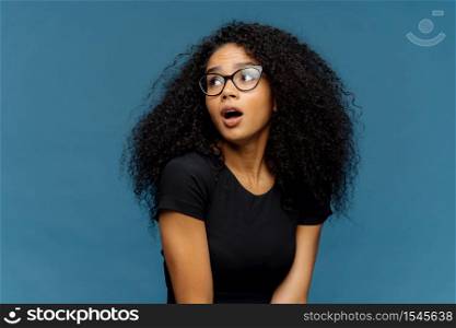 Impressed dark skinned lady with frizzy hair, looks surprisingly aside, keeps jaw dropped, notices something astonishing, wears optical glasses and casual black t shirt, isolated over blue background