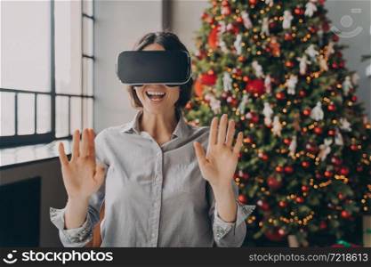 Impressed europian lady using VR googles while spending Christmas at home, playing 3D games in room with beautiful lush Xmas tree on background, happy woman completely immersed in virtual world. Impressed europian lady using VR googles while spending Christmas at home