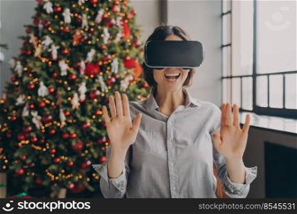 Impressed europian lady using VR googles while spending Christmas at home, playing 3D games in room with beautiful lush Xmas tree on background, happy woman completely immersed in virtual world. Impressed europian lady using VR googles while spending Christmas at home
