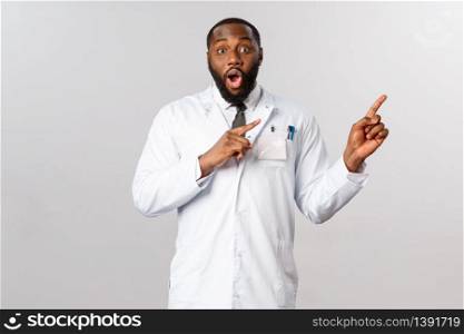 Impressed good-looking male african-american doctor in white coat, pointing upper right corner, gasping and staring camera excited and amazed, tell good news, look its awesome.. Impressed good-looking male african-american doctor in white coat, pointing upper right corner, gasping and staring camera excited and amazed, tell good news, look its awesome