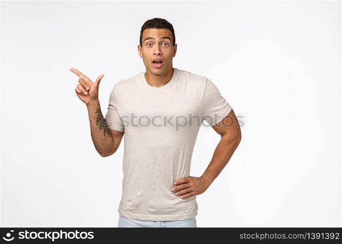 Impressed, surprised handsome tanned man with tattoo on arm, wear casual t-shirt, gasping amazed as discuss new product, pointing left curiously looking camera, want know details about promo.. Impressed, surprised handsome tanned man with tattoo on arm, wear casual t-shirt, gasping amazed as discuss new product, pointing left curiously looking camera, want know details about promo