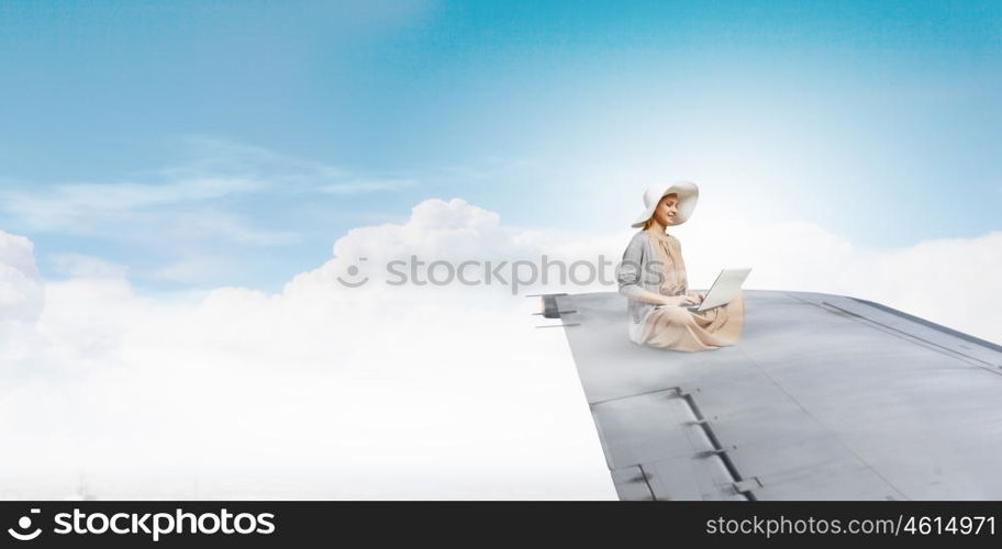 In absolute isolation. Woman in dress and hat sitting on airplane wing and working on laptop