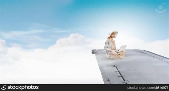 In absolute isolation. Woman in dress and hat sitting on airplane wing and working on laptop