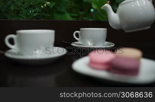 In cafe: pouring tea into two white cups, then focus on hand taking tasty macaroons. Twinkling lights in greenery in background.