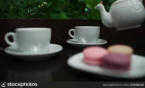 In cafe: pouring tea into two white cups, then focus on hand taking tasty macaroons. Twinkling lights in greenery in background.