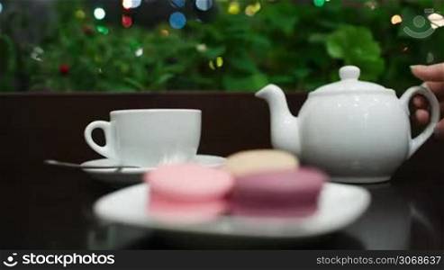 In cafe: pouring tea into white cup, then focus on hand taking tasty macaroons. Twinkling lights in greenery in background.