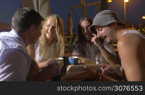 In evening in city of Perea, Greece at the table in cafe sits a young company of 3 men and one girl and they raised their glasses and clinking it with each other. One of the men on tablet shows to other people photo