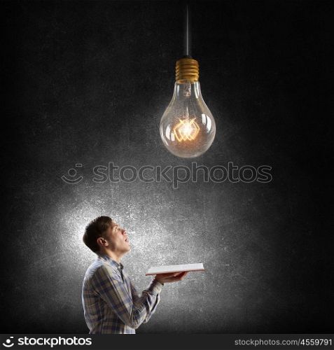 In search of bright inspiration. Young man holding opened book and glass glowing bulb hanging above
